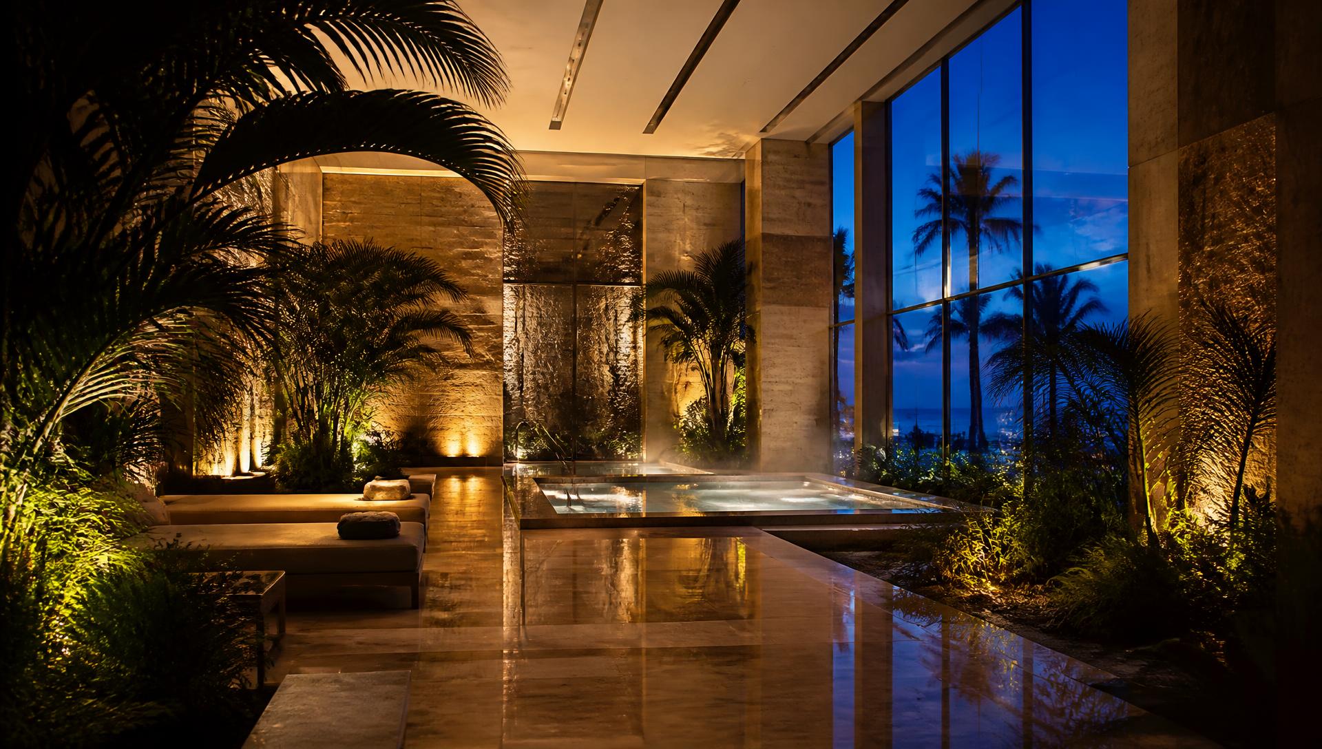 Empty luxury indoor wellness spa at twilight — warm sconce-lit marble, still reflective water and palms against a deep blue evening sky